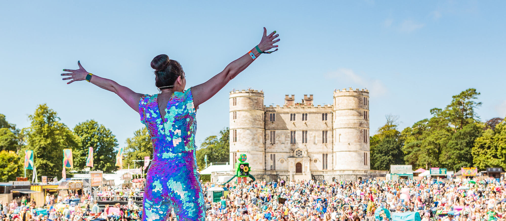 a person in a sequin jumpsuit faces away from the camera looking out on to a crowd of people from the stage. In the distance is a big castle.