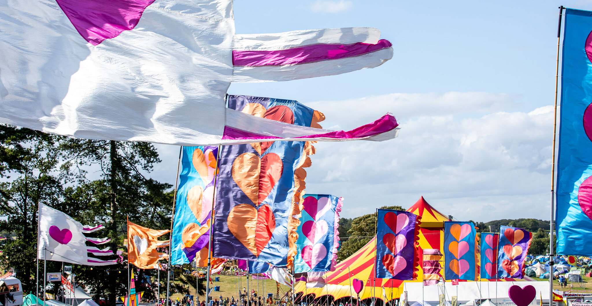 an image featuring multiple giant colourful flags flowing in the wind. Each flag features big hearts. In the distance is a red and yellow circus tent