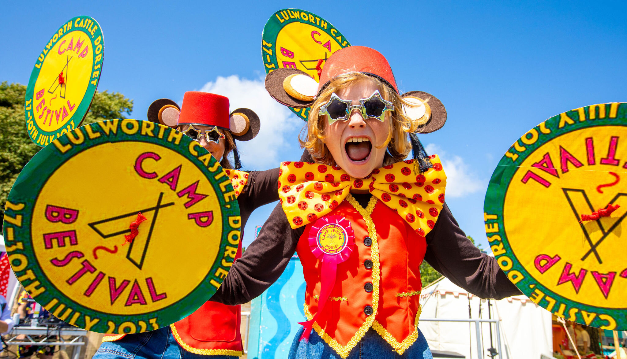 a girl dressed up as a monkey holding symbols shouts with joy in to the camera. She is wearing a red hat, star shaped sun glasses, a giant yellow and red bowtie and a yellow waistcoat.